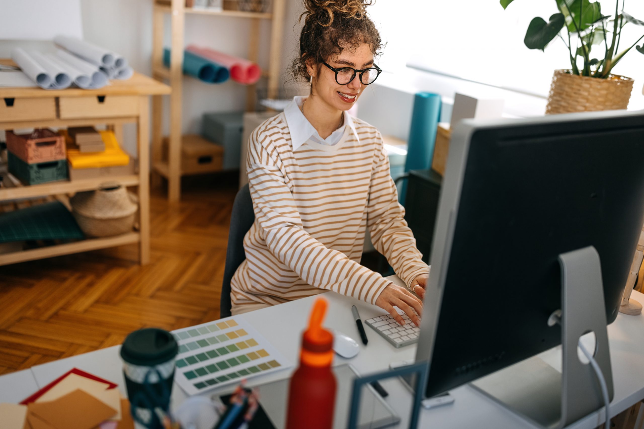 Smiling young woman wearing eyeglasses using desktop computer on desk. She is surrounded by shelves with rolled paper and a cuttered desk.