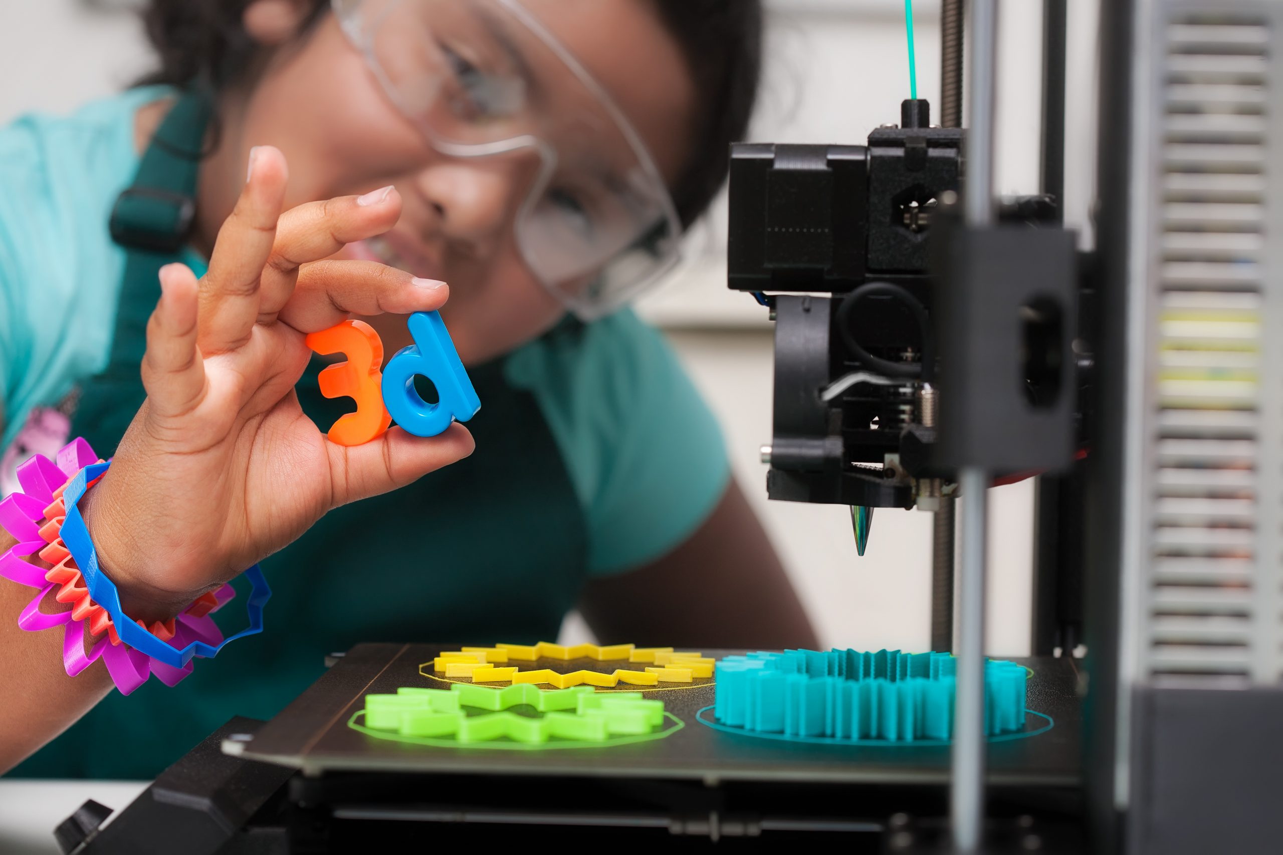 Female student wearing multi-colored 3D printed shapes as bracelets, next to a 3D printer with designs on the heated print bed, and holding 3d letters in her hand.